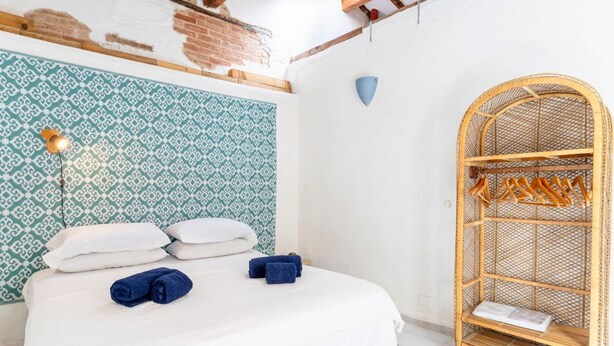 A bed with white linens, a patterned wall, a hanging light fixture, and a wicker shelf with towels.