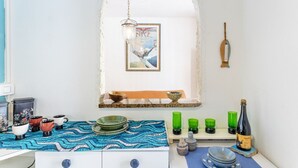 A kitchen with a blue patterned tablecloth, a hanging light fixture, a framed picture on the wall, and a bottle of wine on the counter.