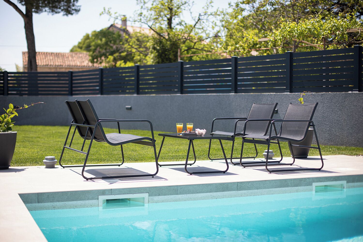 Un área junto a la piscina con dos sillones reclinables, una mesita con una bebida y un plato, y una planta en maceta.