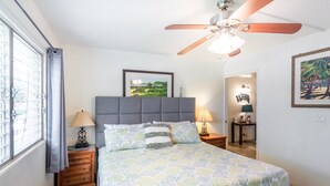A bedroom with a bed, a ceiling fan, a nightstand with a lamp, a framed picture on the wall, and a window with curtains.