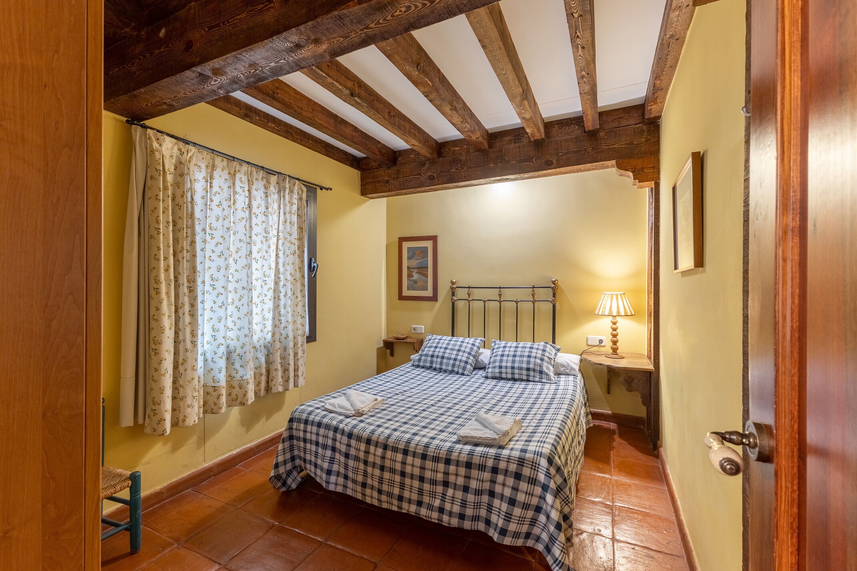 A bedroom with a bed, wooden beams, a nightstand, a lamp, and a window with curtains.
