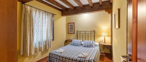 A bedroom with a bed, wooden beams, a nightstand, a lamp, and a window with curtains.