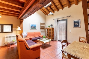 Un salón acogedor con un sofá, una mesa de centro y una mesa de comedor. Hay una ventana con cortinas, un radiador y un techo de madera.
