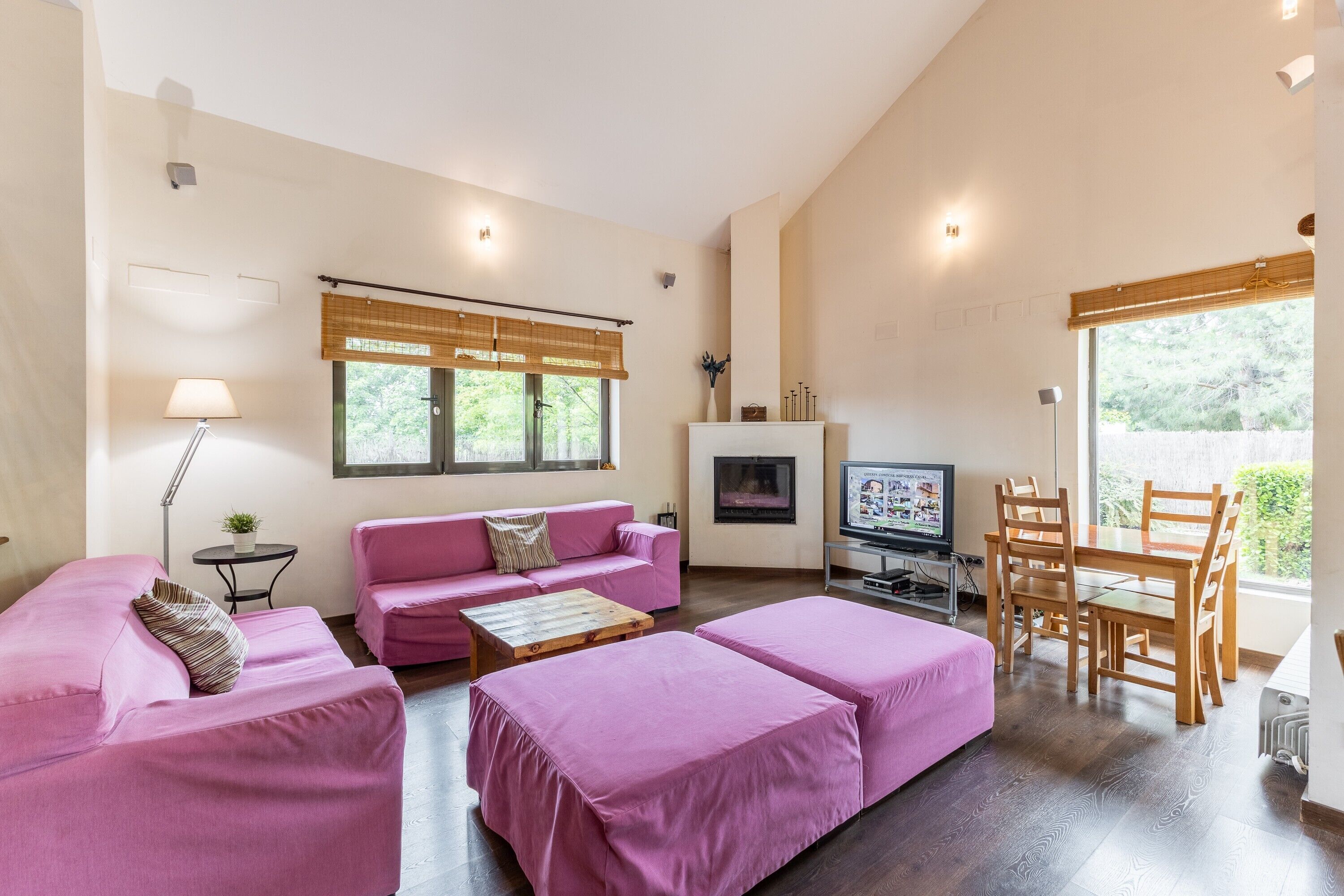 Una habitación con un sofá rosa, una mesa de comedor de madera, un televisor y una ventana con vistas a zonas verdes.