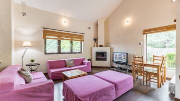 Una habitación con un sofá rosa, una mesa de comedor de madera, un televisor y una ventana con vistas a zonas verdes.