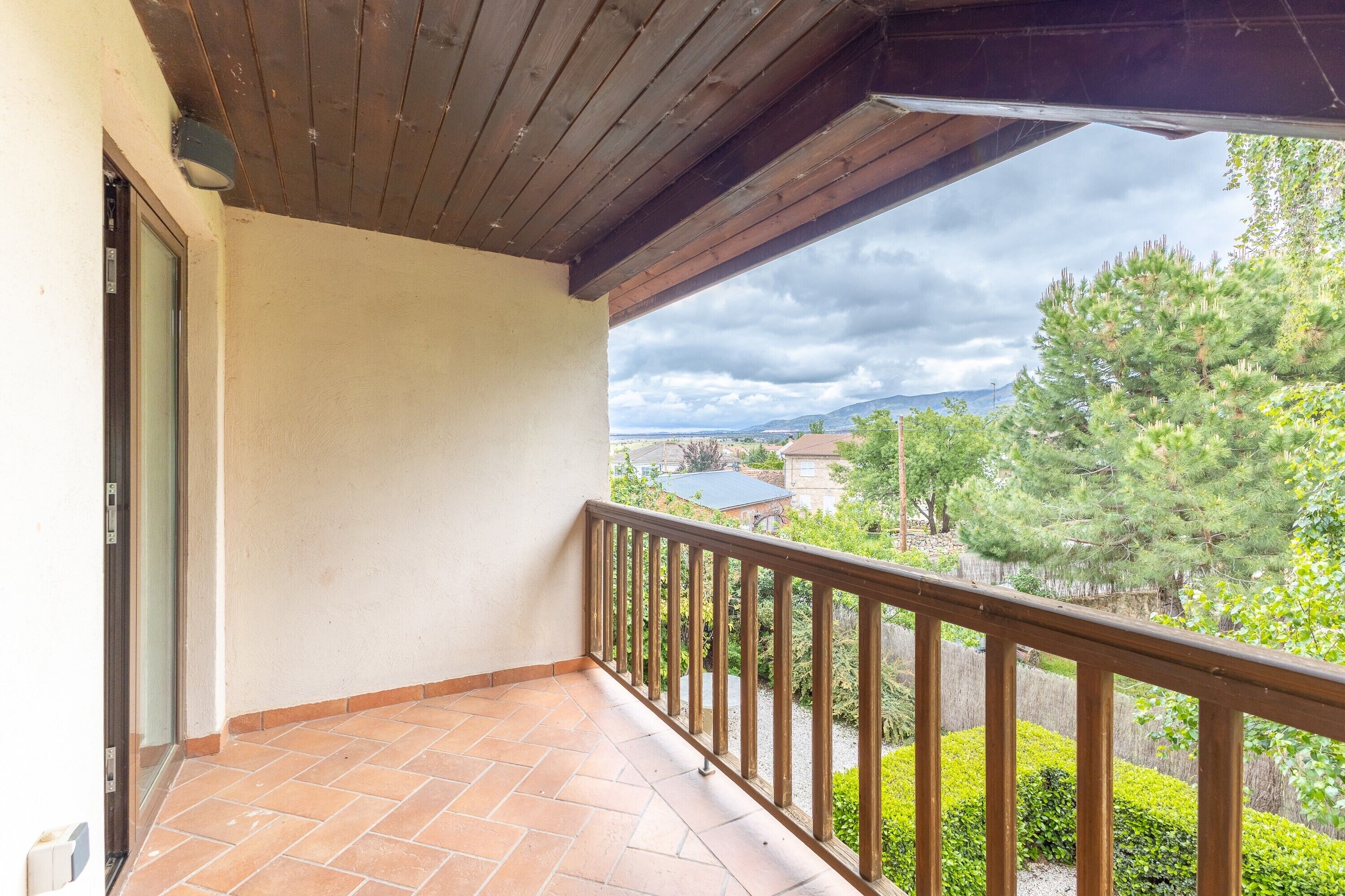 A balcony with a wooden railing, a tiled floor, and a view of trees and buildings in the distance.