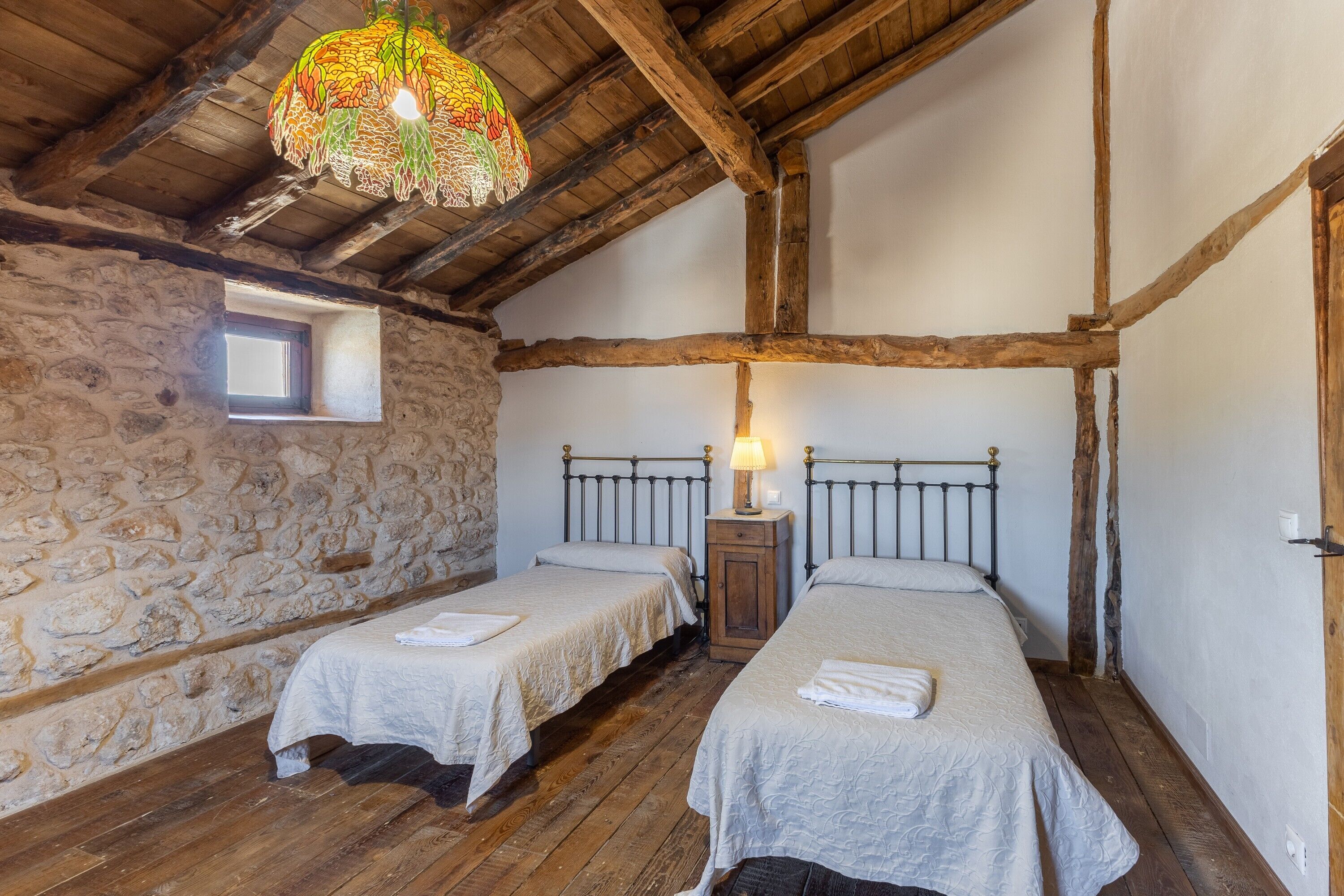 Een rustieke kamer met twee bedden, een stenen muur, houten balken en een hanglamp.