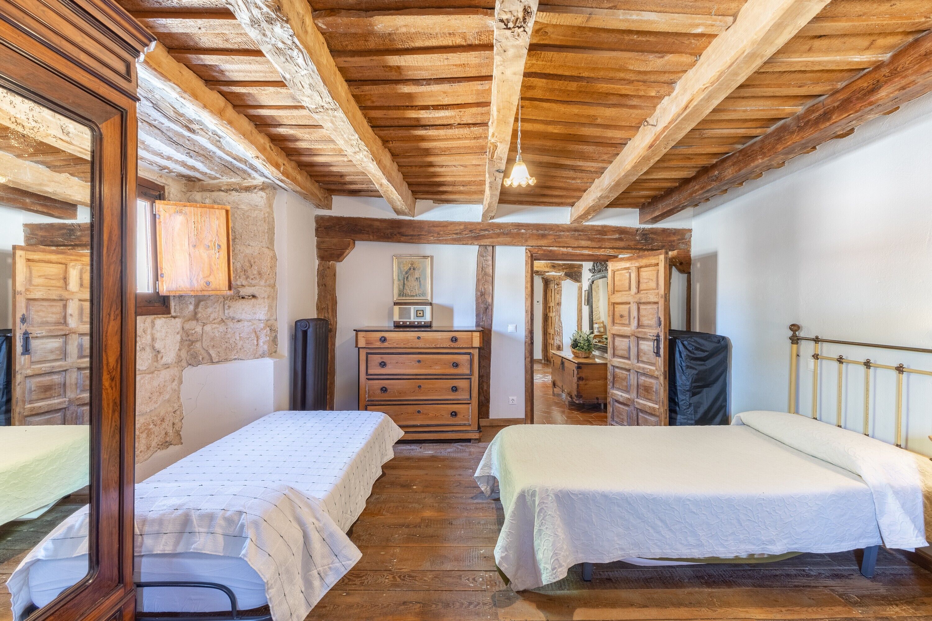A rustic room with wooden beams, a stone wall, a dresser, two beds, and a mirror.