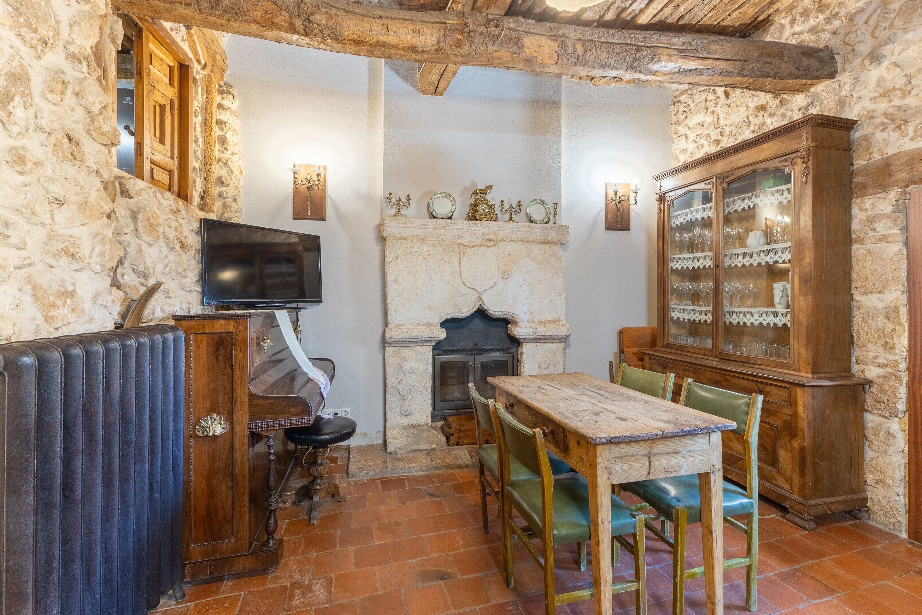 Una habitación rústica con chimenea, paredes de piedra, una mesa de madera con sillas, un aparador con puertas de cristal y un televisor.