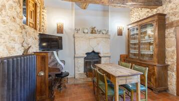 Una habitación rústica con chimenea, paredes de piedra, una mesa de madera con sillas, un aparador con puertas de cristal y un televisor.