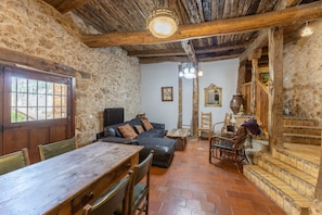 A rustic interior with stone walls, a wooden ceiling, a dining table, a sofa, and a staircase.