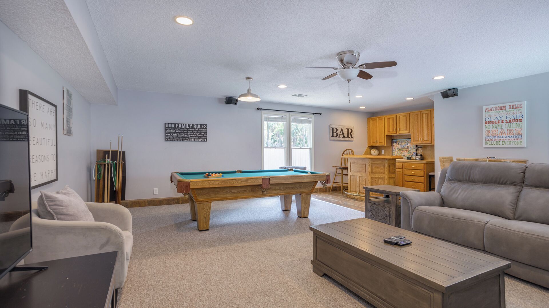 A living room with a pool table, two sofas, a coffee table, and a kitchen area with wooden cabinets.