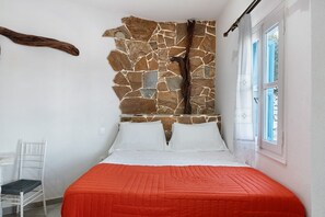 A bedroom with a stone wall, a bed with white and orange bedding, a chair, and a window with curtains. - Studio Apartment 'Politimi  N1' with Mountain View, Wi-Fi and Air Conditioning (Amorgos)