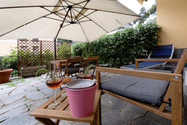 En patio med et træbord, en lyserød spand og et glas vin under en stor parasol.