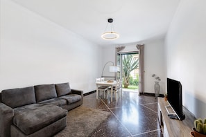A modern living room with a grey sofa, a dining table with chairs, a flat-screen TV, and a large window with a view of greenery.