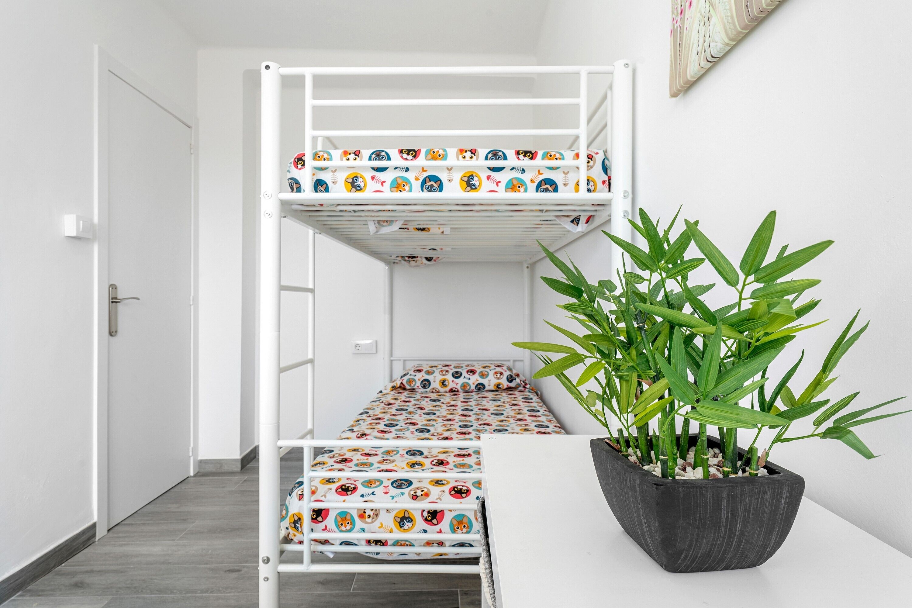 A bunk bed with a colorful panda-themed mattress, a potted plant, and a white wall with a framed picture.