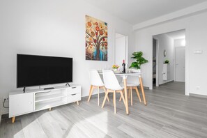A modern living room with a flat-screen TV, a dining area with white chairs and a table, and a hallway leading to another room.