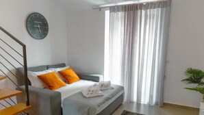 Un dormitorio moderno con un sofá gris, almohadas naranjas, una manta blanca, un reloj, una escalera y una planta.