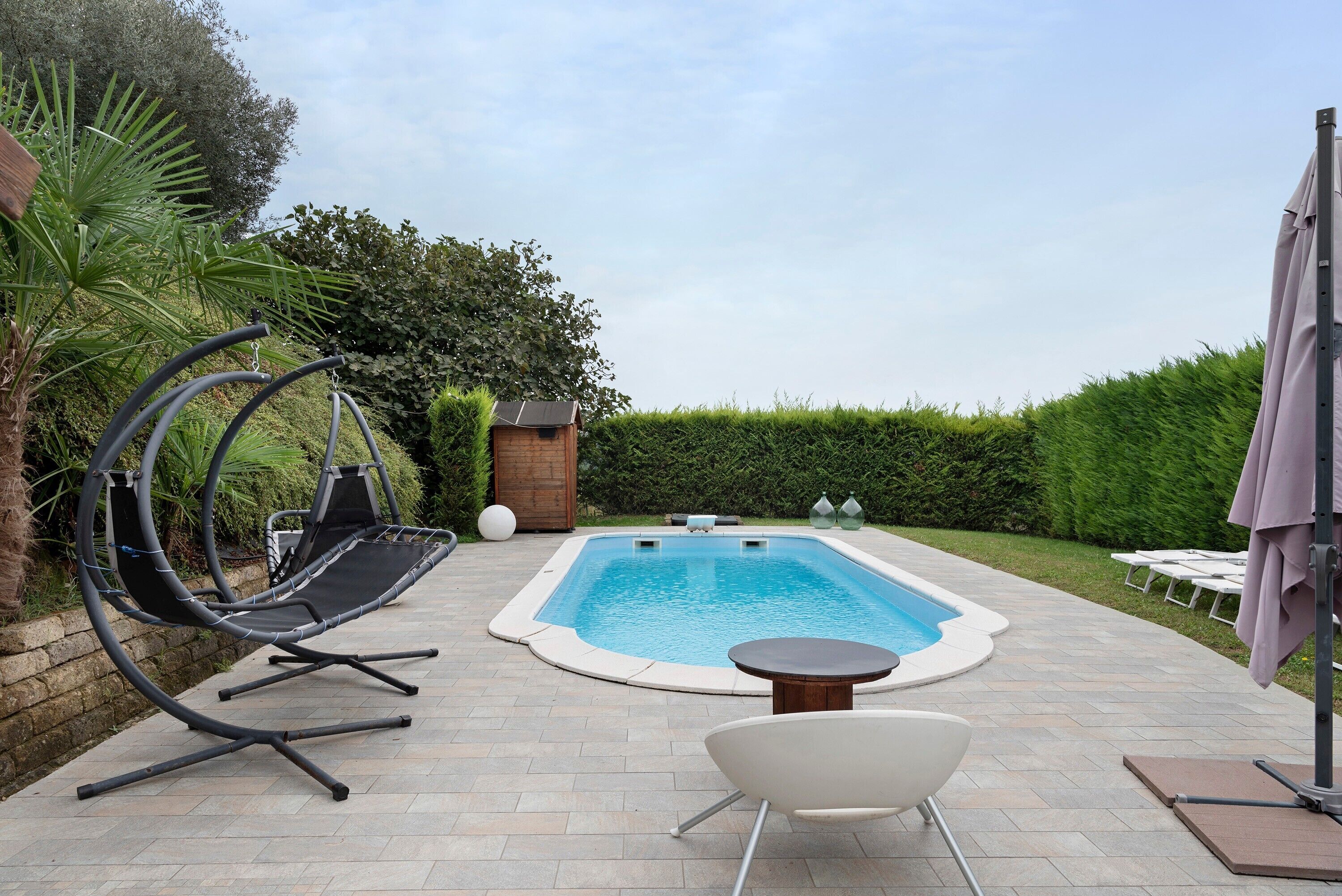 Una piscina rectangular con un bordeado de baldosas, una silla colgante, una tumbona blanca y una pequeña mesa.