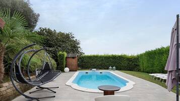 Una piscina rectangular con un bordeado de baldosas, una silla colgante, una tumbona blanca y una pequeña mesa.