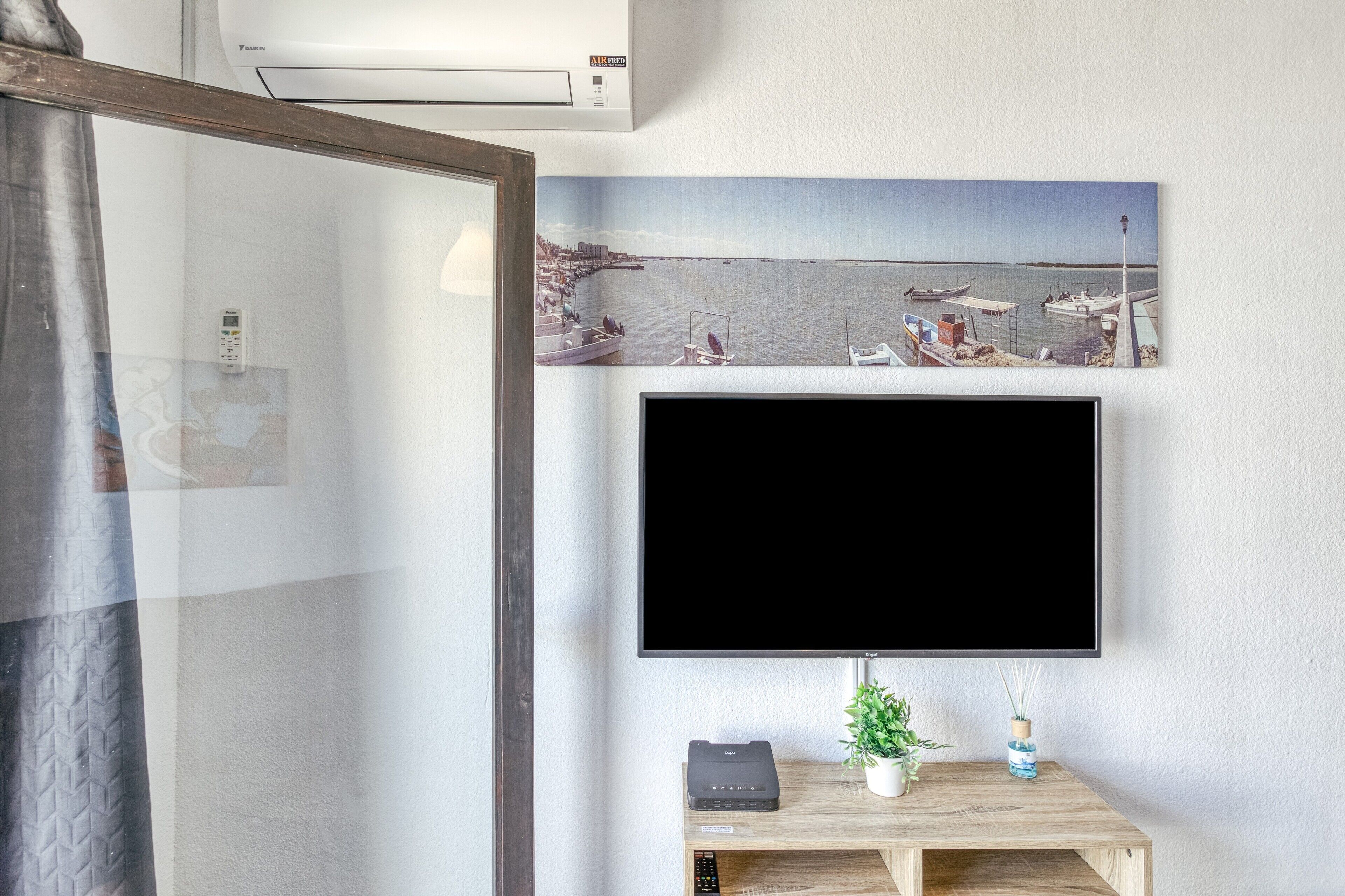 A room with a mounted TV, a wooden side table with a plant, and a framed picture of a beach scene.