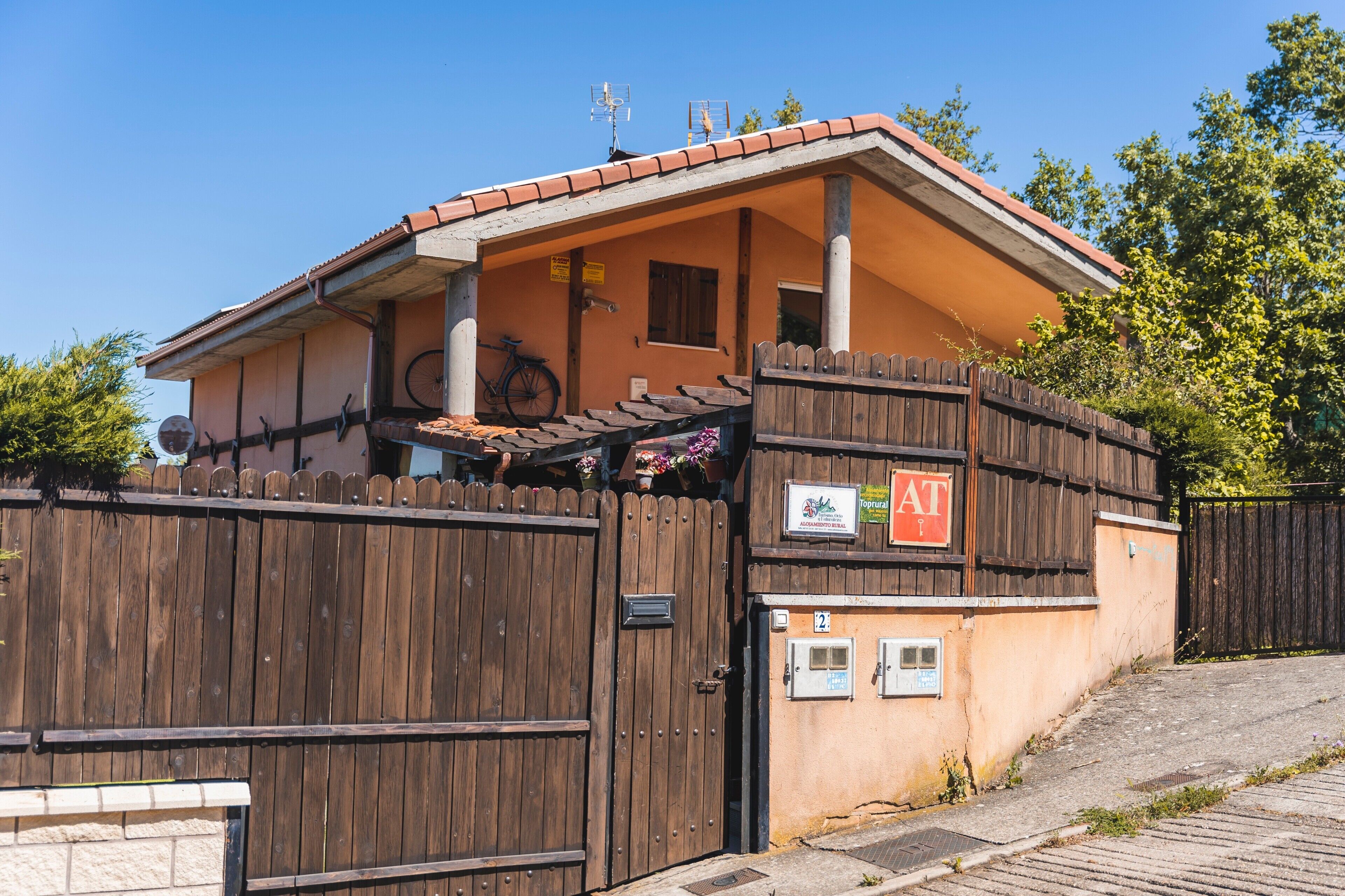Un bâtiment à deux étages, avec une clôture en bois, une enseigne indiquant « AT » et un vélo garé à l’extérieur.