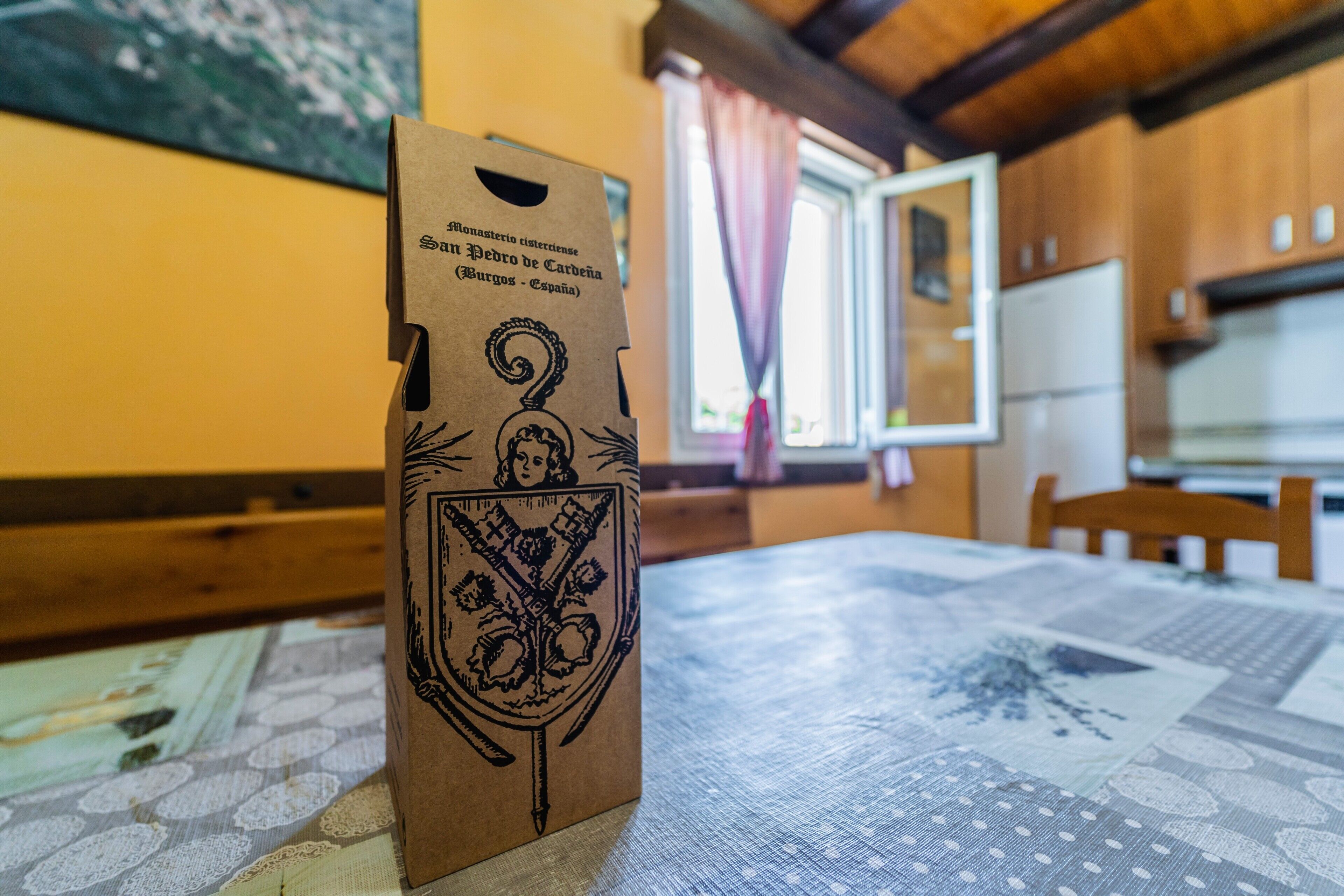 A table with a patterned tablecloth, a brown paper bag with a crest, and a window with curtains in the background.