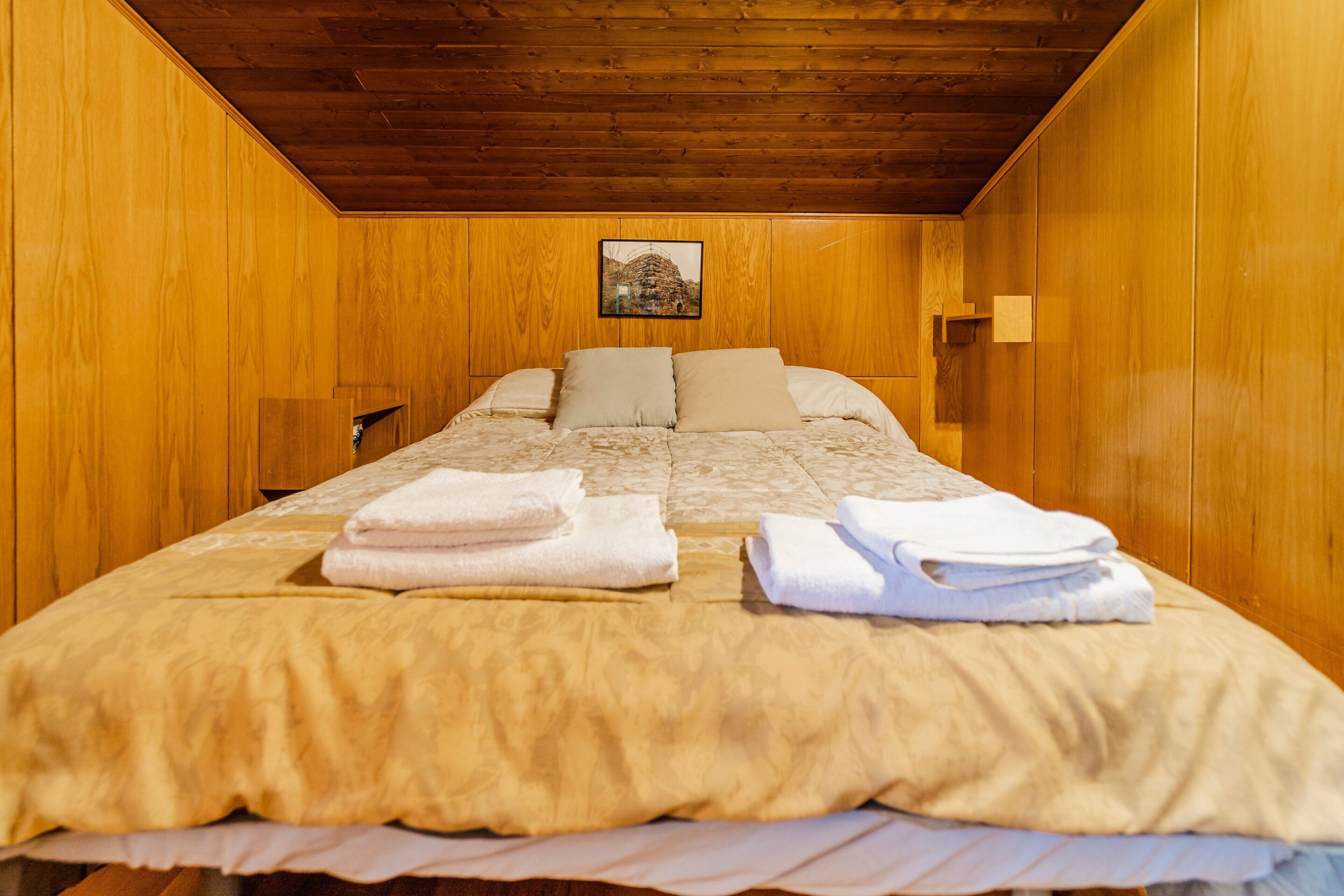 Habitació amb parets de fusta, un llit amb edredó beix, dos coixins i tovalloles doblegades.