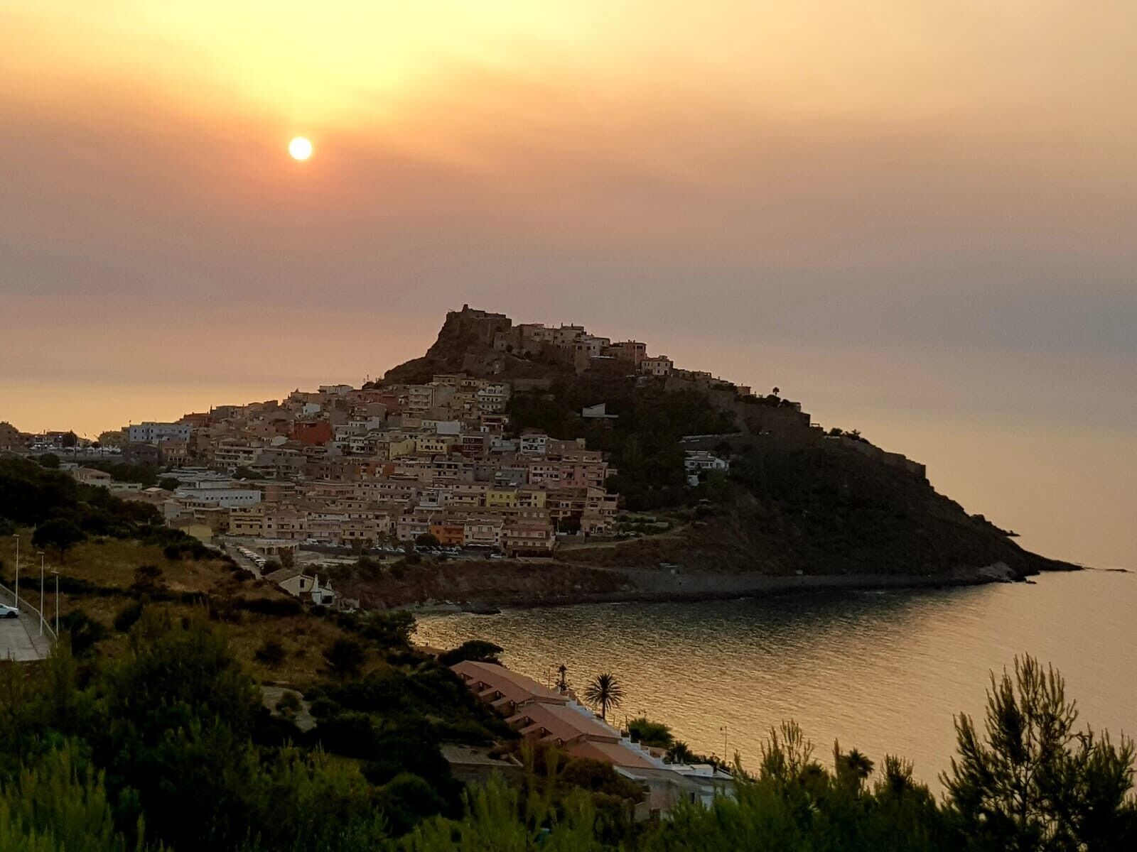 A coastal town with buildings on a hill, a body of water, and a sunset.