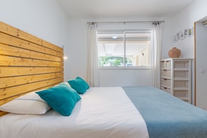 A bedroom with a wooden headboard, a bed with white and blue bedding, a window with white curtains, and a white shelving unit with baskets.