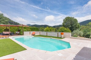 Ein rechteckiger Swimmingpool mit einer gefliesten Terrasse, umgeben von einer Rasenfläche und einer Pergola mit Gartenmöbeln.