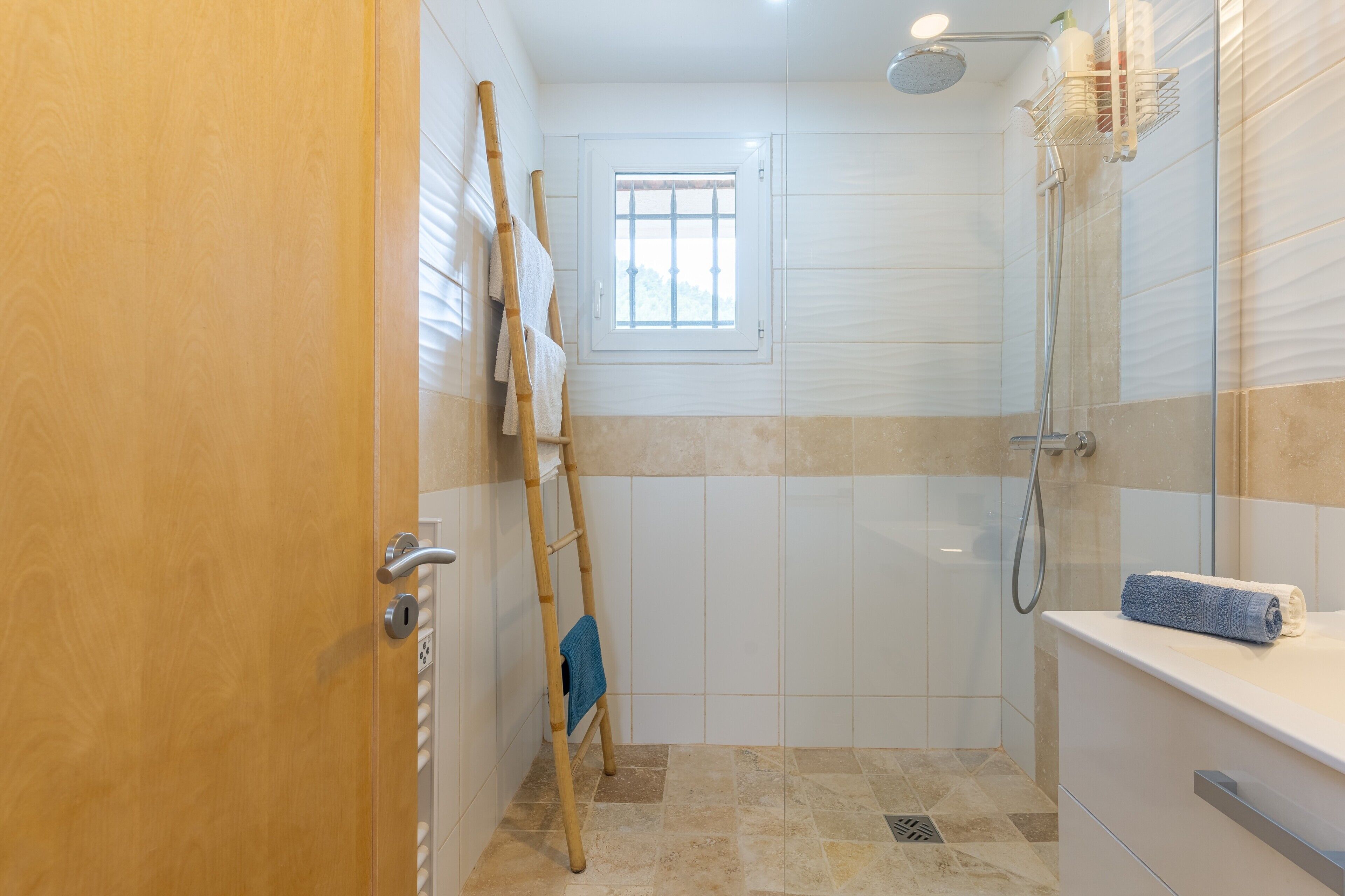 A bathroom with a shower, a window, a towel rack, and a wooden door.