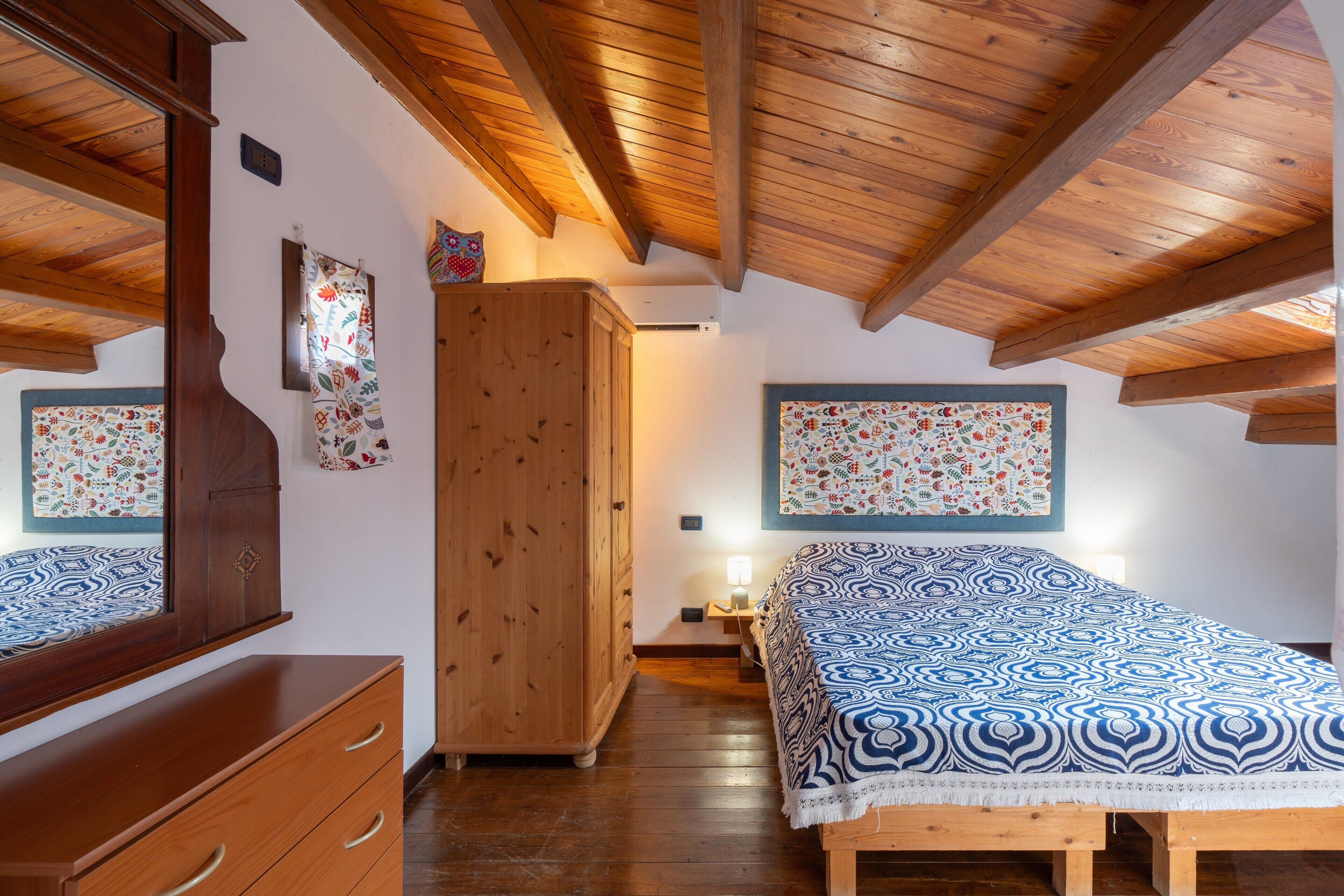 A bedroom with a wooden ceiling, a bed with patterned bedding, a wardrobe, and a mirror.