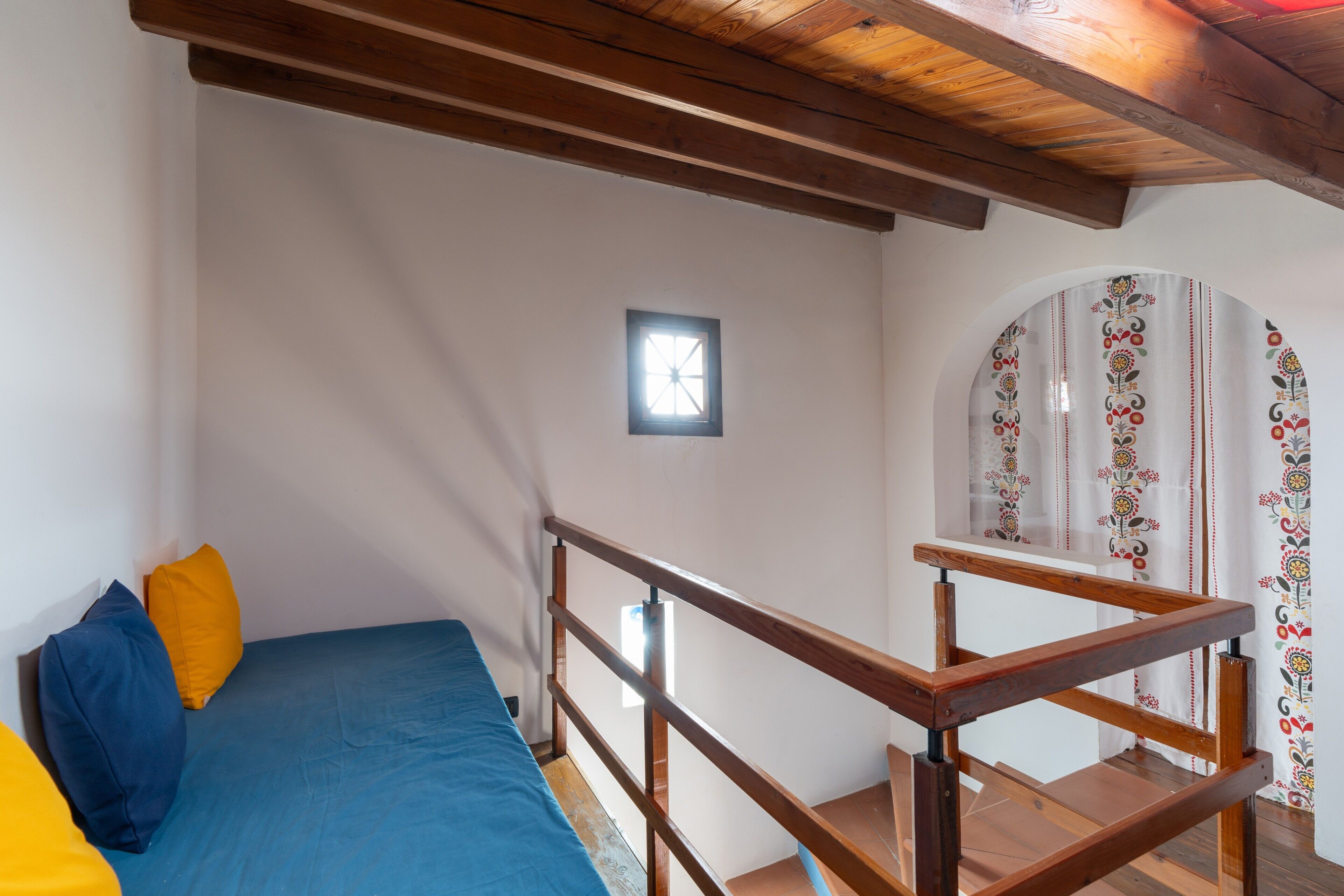 Una habitación con cama, vigas de madera en el techo, una ventana pequeña y una cortina estampada.