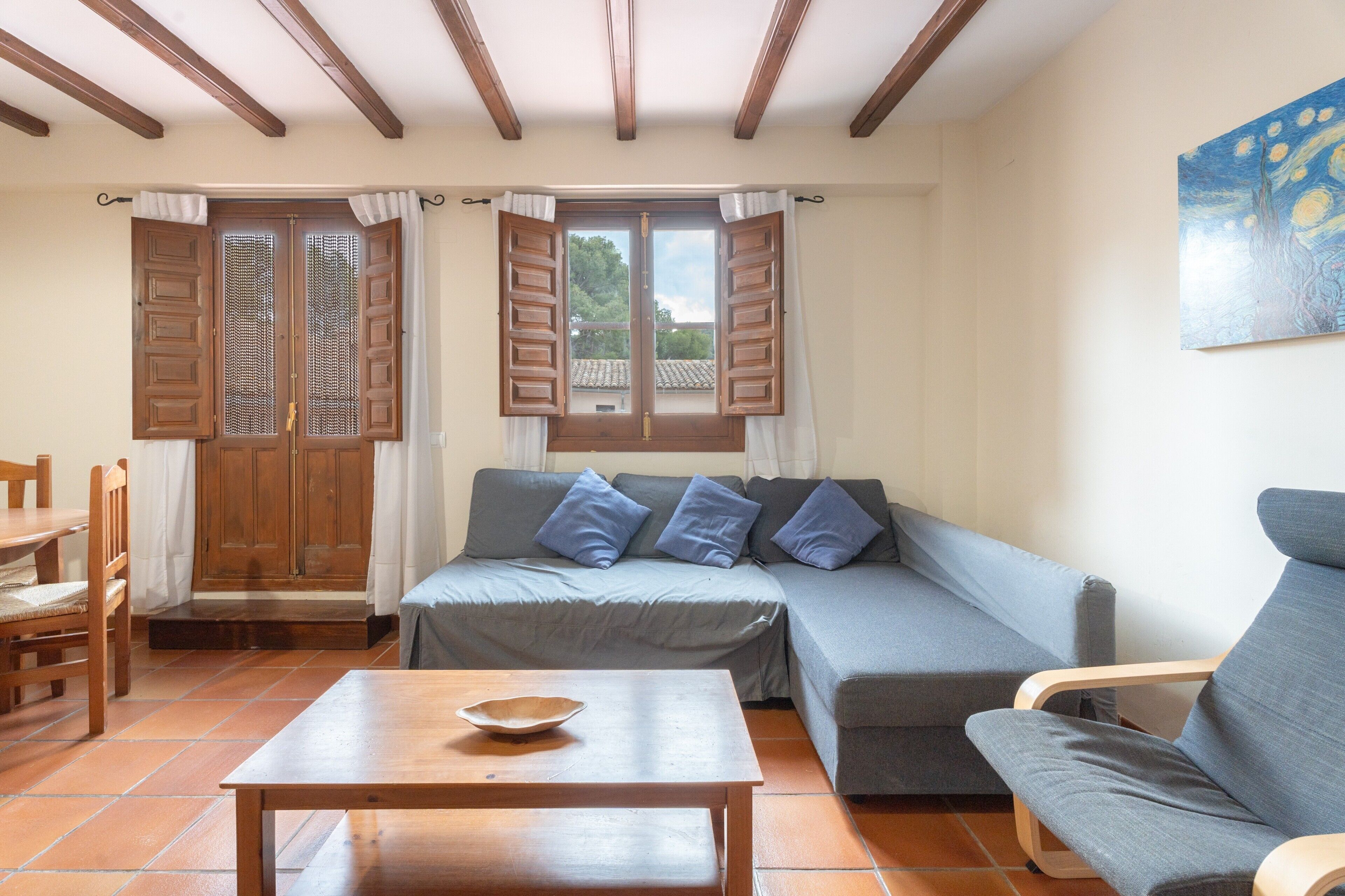 A living room with a sofa, a wooden table, and a chair. There are two windows with wooden shutters, and a painting is visible on the wall.