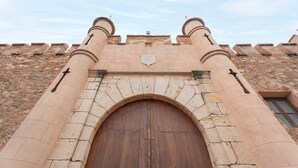 Un arco de piedra con una puerta de madera, flanqueado por dos torres cilíndricas con cruces, que conduce a un edificio de fachada de piedra.