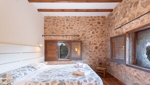 A bedroom with a stone wall, wooden beams, a bed with floral bedding, a window with shutters, and a chair. - Apartment 'Bruno' with Mountain View, Shared Pool and Shared Garden (Altura)