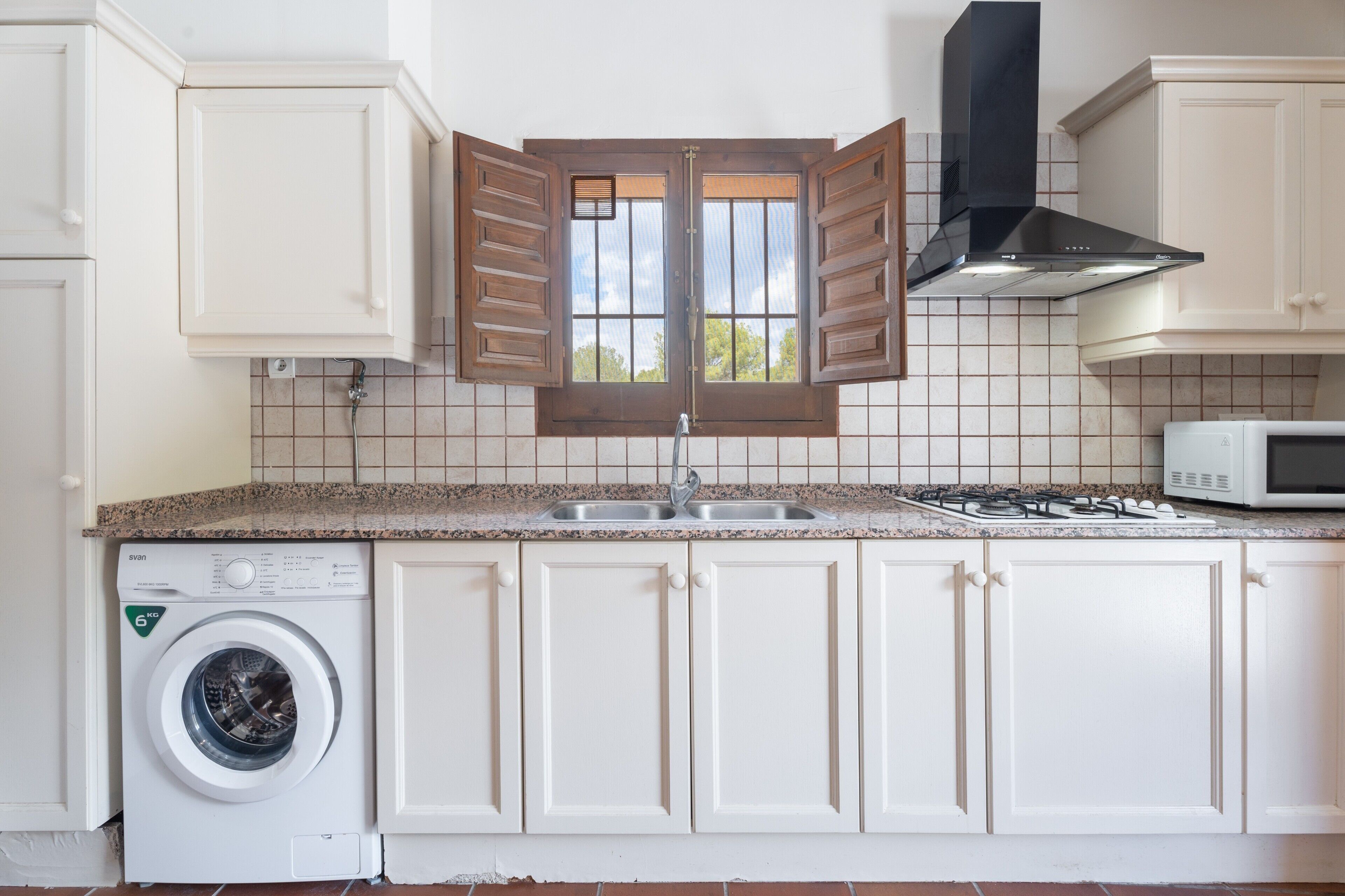 Una cucina con mobili bianchi, un forno a microonde integrato, un piano cottura a gas, una lavatrice e una finestra con persiane di legno.
