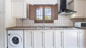 Una cucina con mobili bianchi, un forno a microonde integrato, un piano cottura a gas, una lavatrice e una finestra con persiane di legno.
