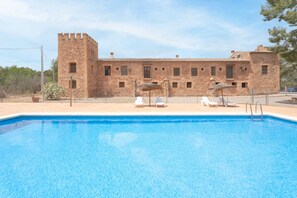 Une piscine avec des chaises longues, un bâtiment en pierre doté d’une tour et un ciel dégagé.
