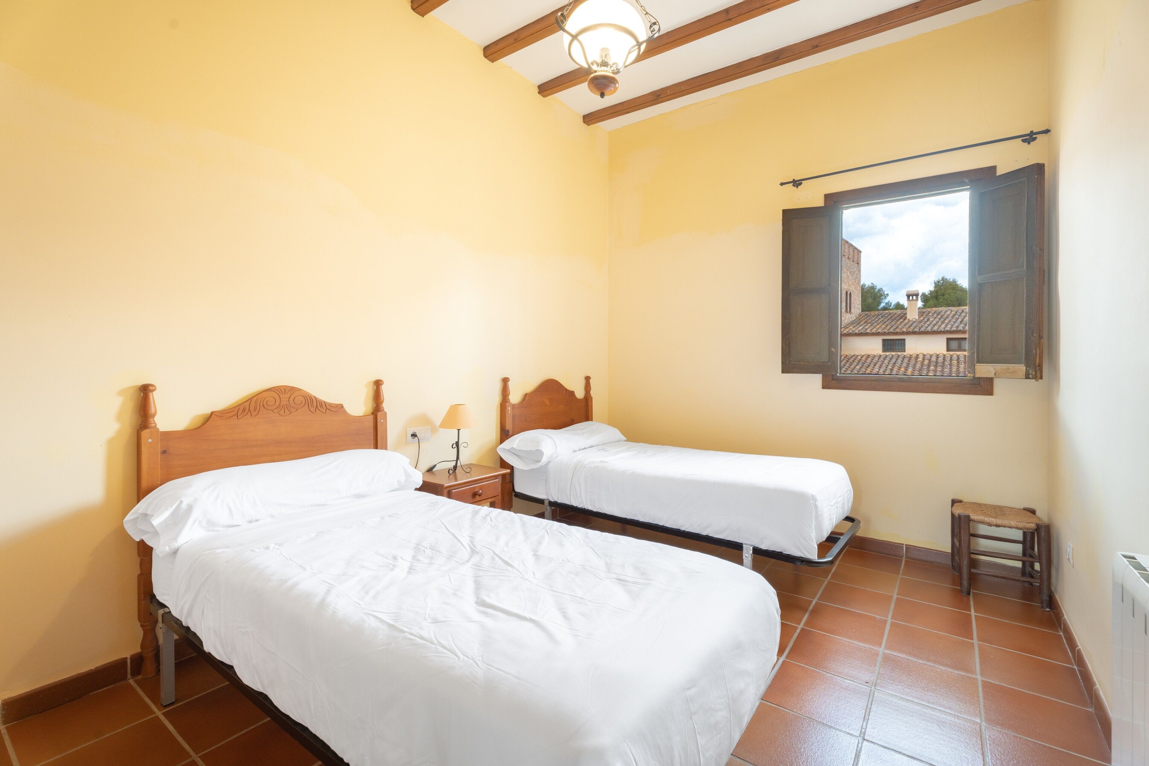 Une chambre avec deux lits simples, des têtes de lit en bois, une petite table de chevet avec une lampe, une fenêtre donnant sur des immeubles et un radiateur au mur.