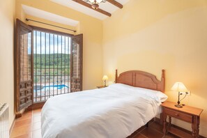 Un dormitorio con una cama grande, una mesita de noche de madera con una lámpara, y vista a una piscina por una puerta abierta.