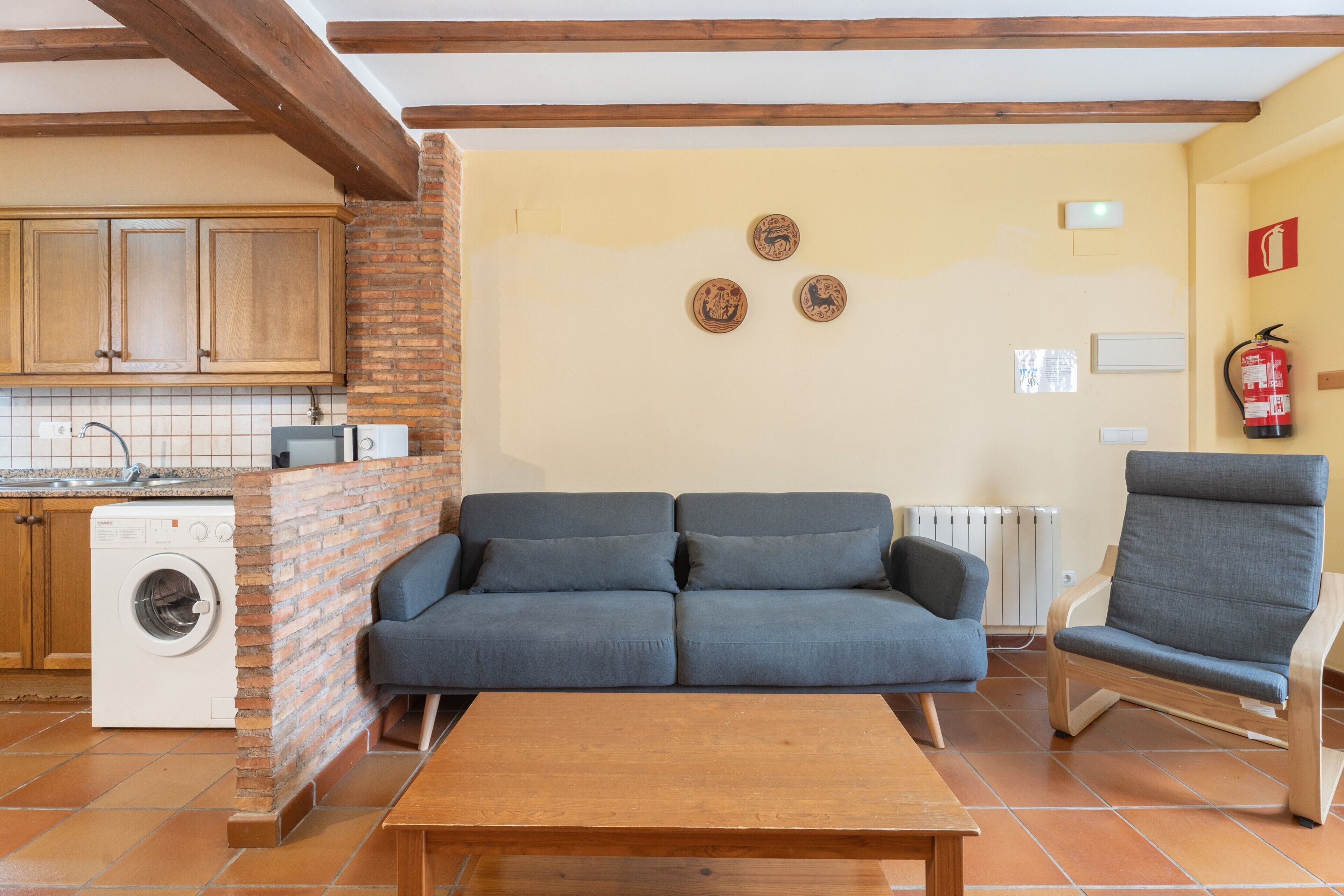 Een woonkamer met een bank, een stoel, een houten tafel, een wasmachine en een bakstenen muur.