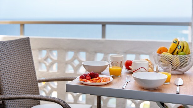 Una mesa de desayuno con fruta, jugo y un bol en un balcón con vista al mar.