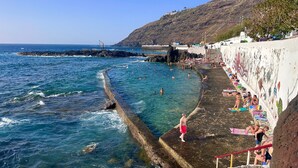 Escena costera con un litoral rocoso, agua azul cristalina y una playa donde personas nadan y toman el sol.