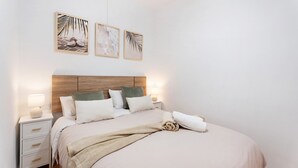 Un dormitorio ordenado con cabecera de madera, una mesita de noche blanca con lámpara, y una cama con manta color crema.