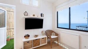 Una sala de estar con televisor de pantalla plana, muebles de mimbre, un pequeño estante con adornos y vista al mar a través de un ventanal grande.