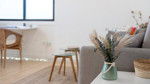 Una sala de estar con un sofá, una mesita redonda pequeña, una cesta de mimbre y un florero con plantas secas.