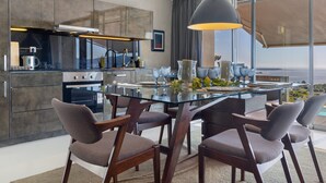 Une cuisine moderne avec une table à manger en verre, des chaises foncées et un grand luminaire suspendu.