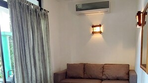 Quarto com ventoinha no teto, sofá, mesa pequena, janela com cortinas e ar condicionado fixado na parede.